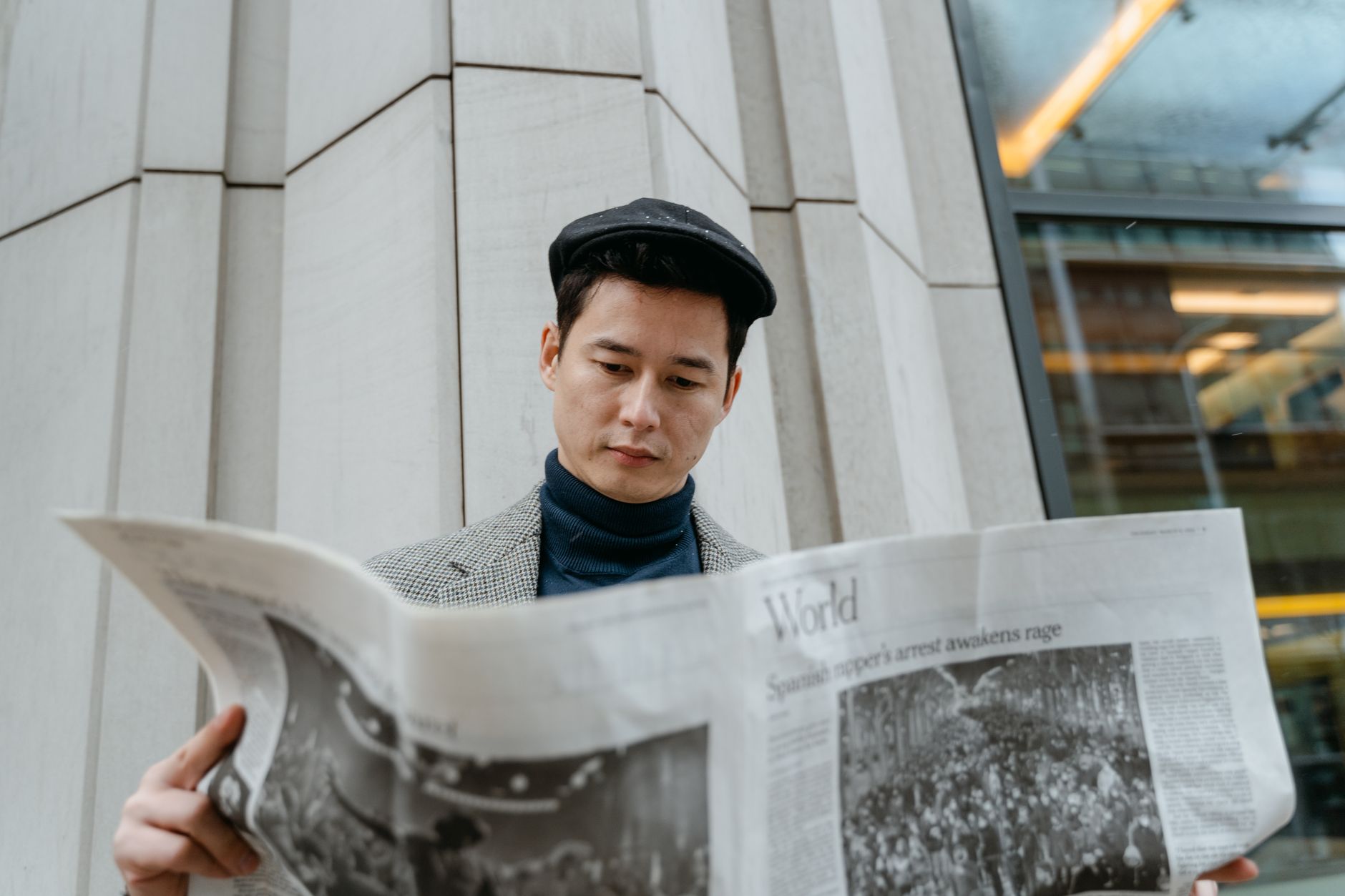 A young man in a flat cap and turtleneck reads a newspaper outdoors. — A Guide to Monetizing Your Selfies