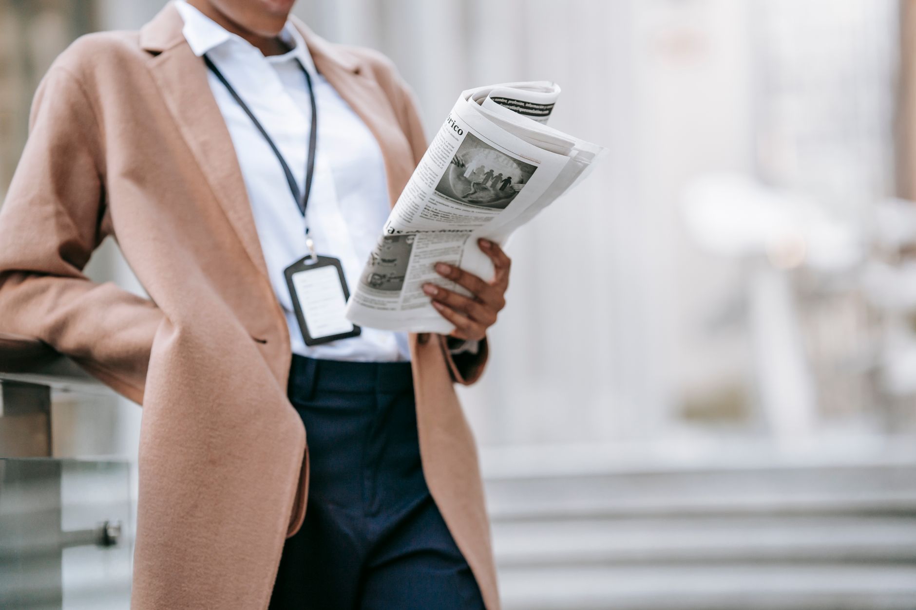Crop unrecognizable young African American female entrepreneur in elegant brown coat with badge reading newspaper while leani