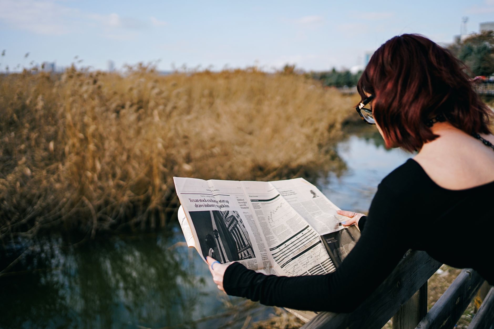 A woman reads a newspaper by a river, surrounded by autumn reeds and tranquil scenery. — Selling Your Essays For Cash
